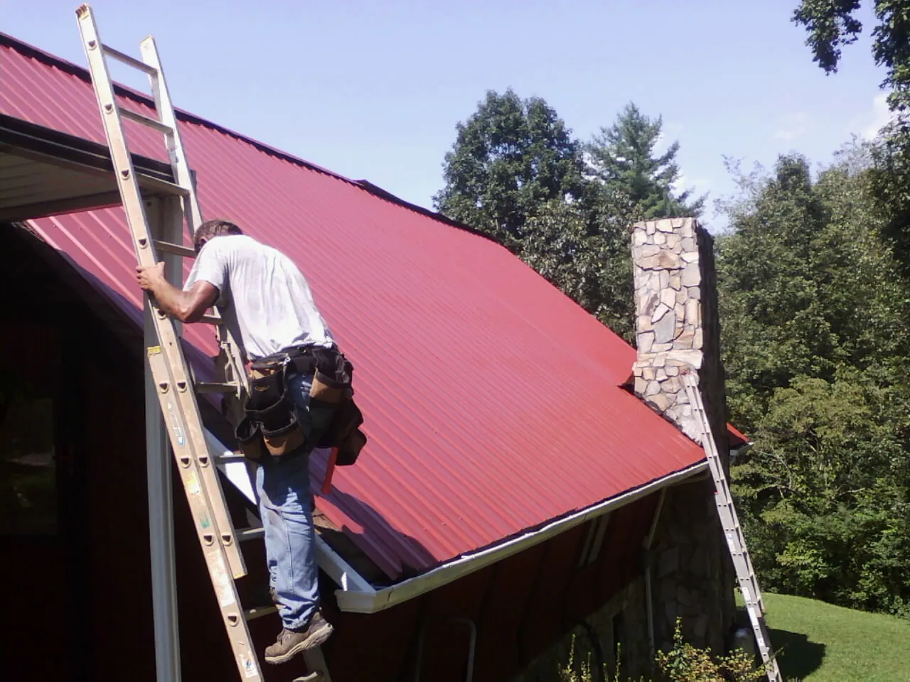 Roofer on a ladder beside a red metal roof and stone chimney — Soffit & Fascia Repair by Roofing Brothers in Pickens